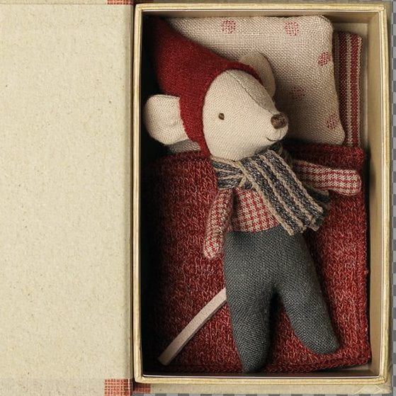 A small plush mouse toy dressed in a red hat and scarf with gray pants, nestled inside an open beige fabric gift box with a red patterned pillow.