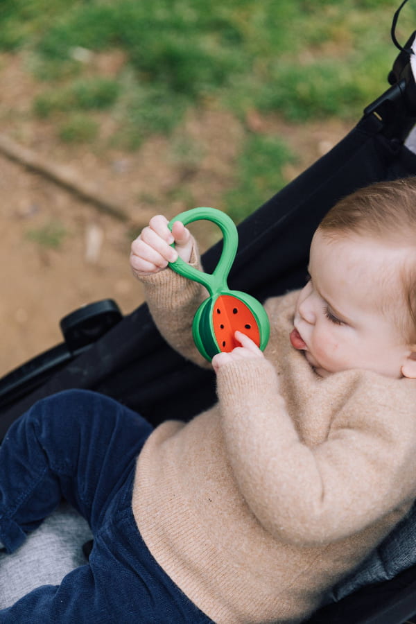 Baby holding a green teething ring with a watermelon design in a stroller.