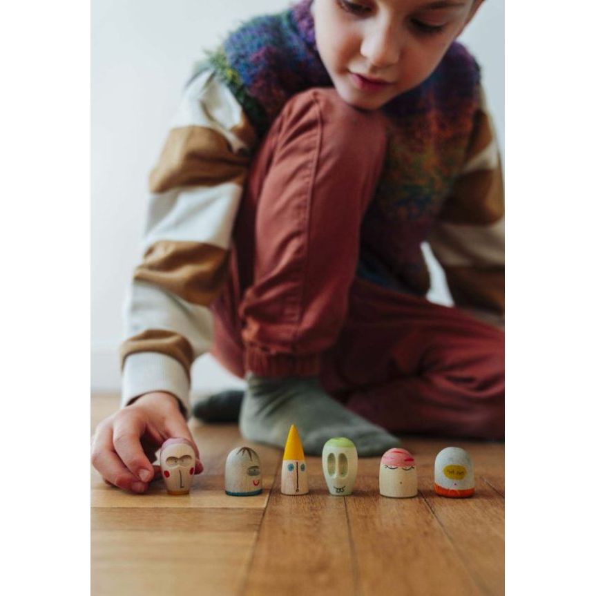 A child wearing a colorful knit sweater and rust-colored pants sits on a wooden floor, reaching for a small wooden toy shaped like a face. Five other small wooden toys are lined up in front of the child.