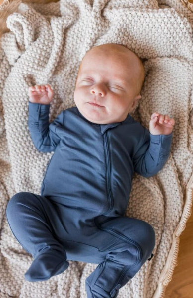 A sleeping newborn baby wearing a navy blue footed zip-up onesie, lying on a cozy beige knit blanket.