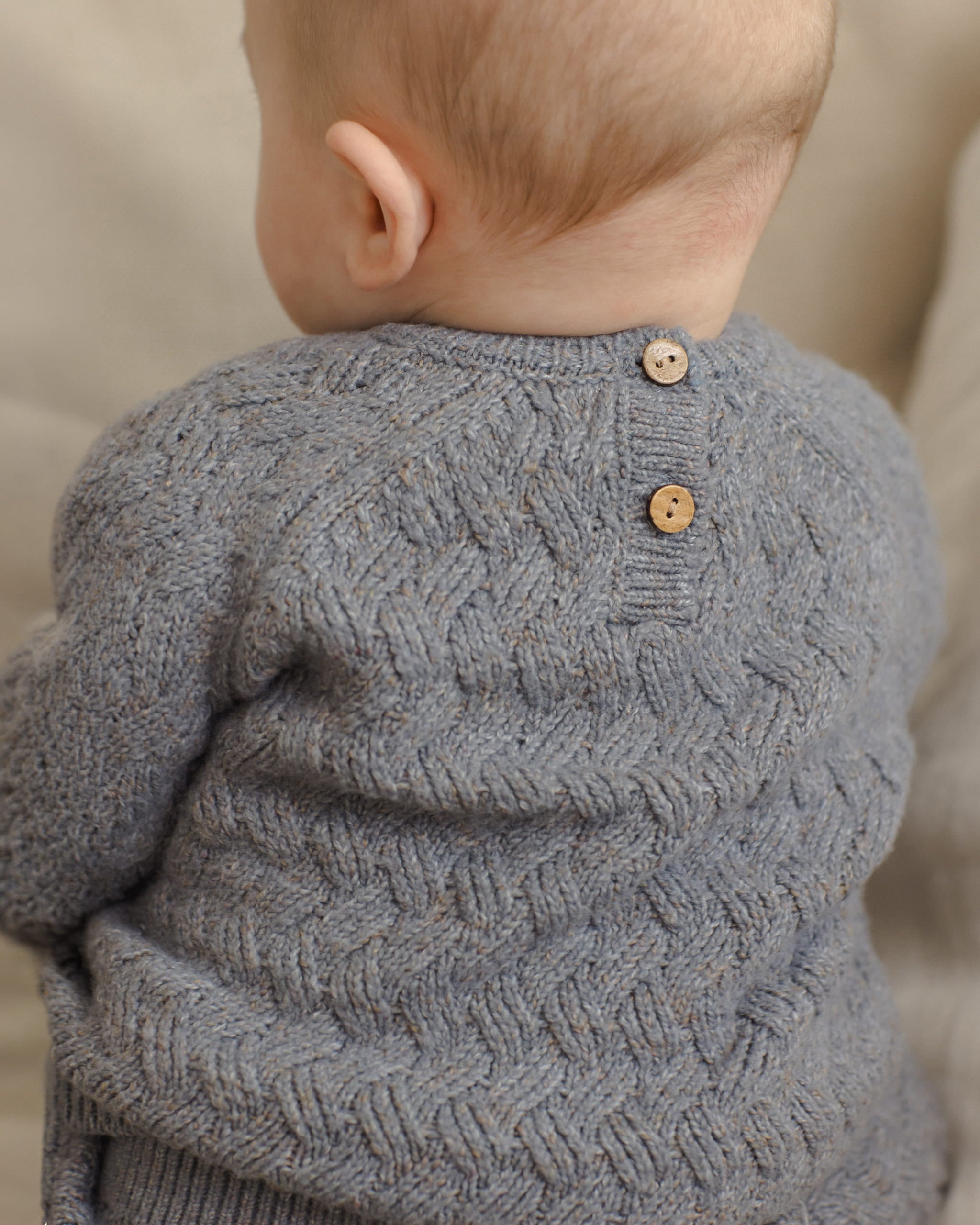 Close-up back view of a baby wearing a gray textured knit sweater with two wooden buttons at the neckline, set against a soft beige background.