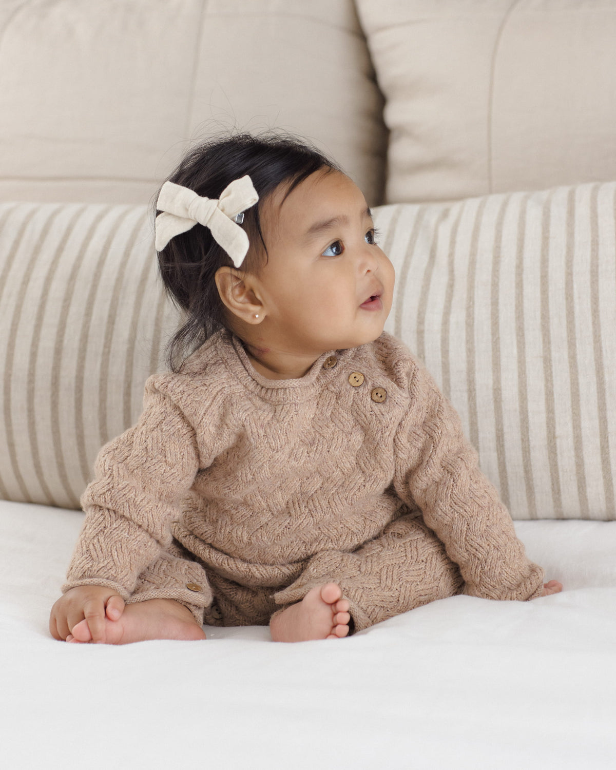 A baby wearing a beige cable-knit romper and matching hat, sitting on a white blanket in front of striped pillows.