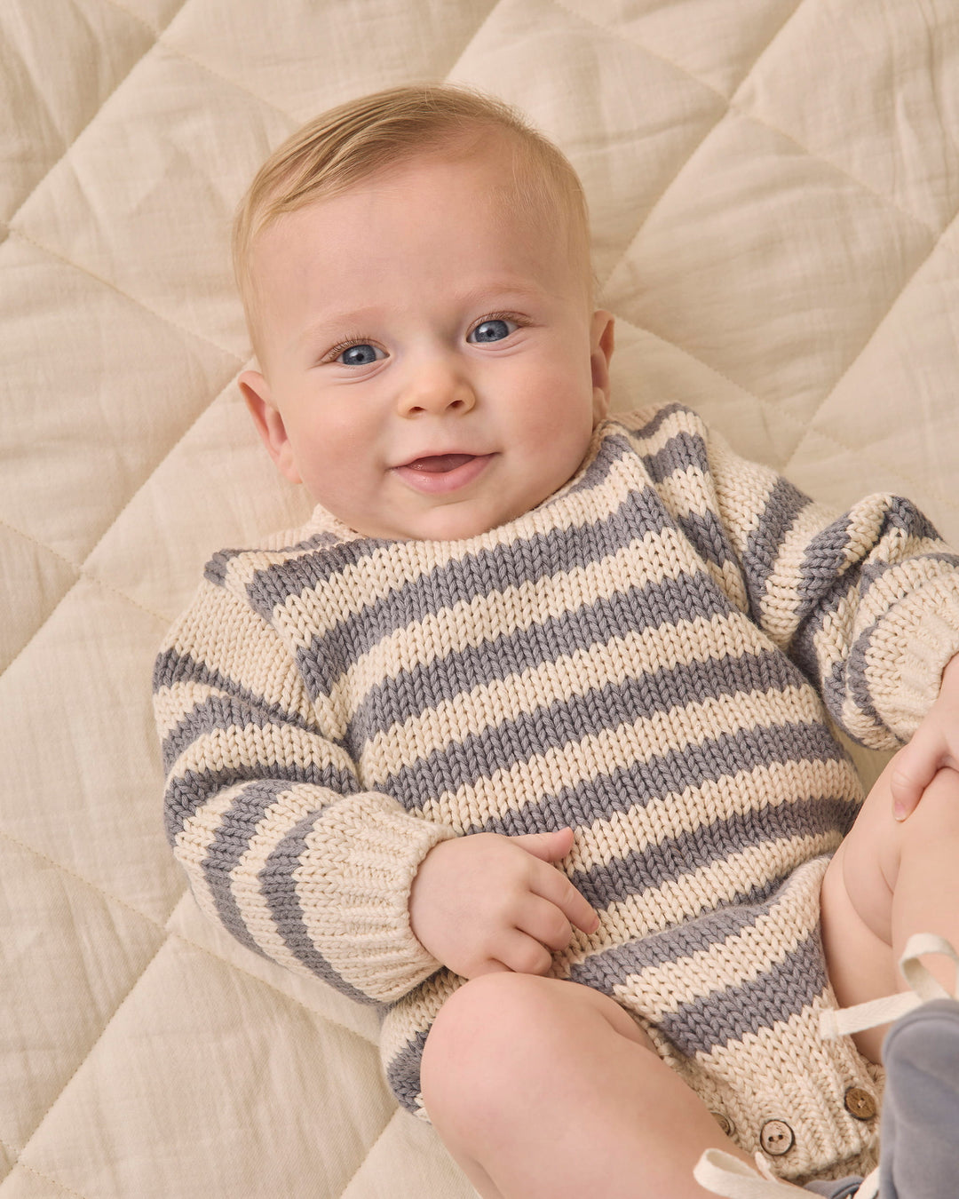 A baby lying on a quilted cream blanket, wearing a cream and gray striped knit long-sleeve romper.