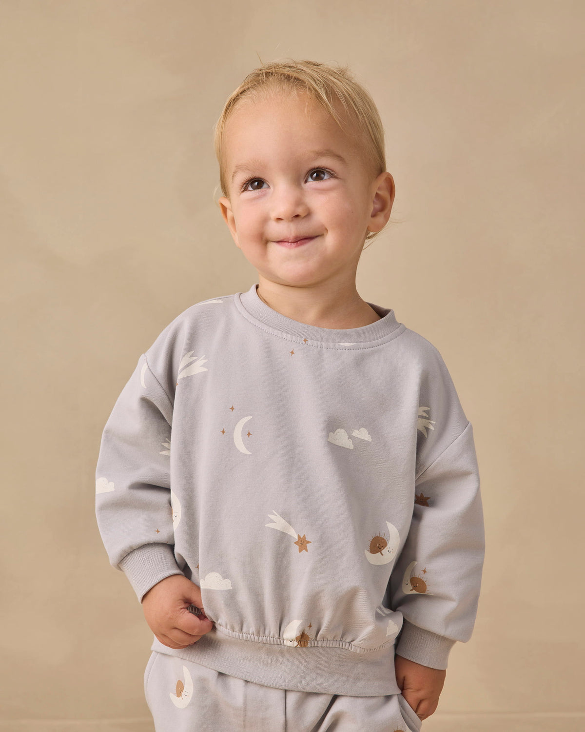 A smiling toddler with blonde hair wearing a light gray long-sleeve sweatshirt and matching pants with a celestial print of white clouds, moons, and stars, set against a plain beige background.