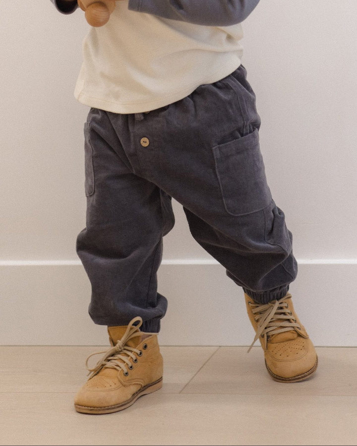 A toddler standing indoors against a plain light wall, wearing a cream and navy long-sleeve henley top, dark charcoal pants, and tan lace-up boots, while holding a small wooden toy.