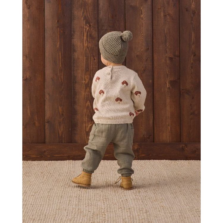 A toddler standing with their back to the camera, wearing a cream knit sweater with small brown motifs, a gray knit beanie with a pom-pom, olive green pants, and tan boots, set against a dark wood plank wall and a light woven rug.