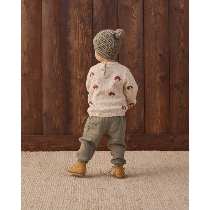 A toddler standing with their back to the camera, wearing a cream knit sweater with small brown motifs, a gray knit beanie with a pom-pom, olive green pants, and tan boots, set against a dark wood plank wall and a light woven rug.