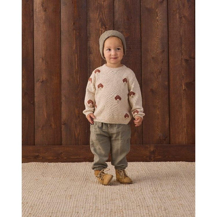 A toddler stands in front of a dark wood plank wall wearing a cream knit sweater with small brown tree motifs, olive green pants, tan boots, and a beige knit beanie.