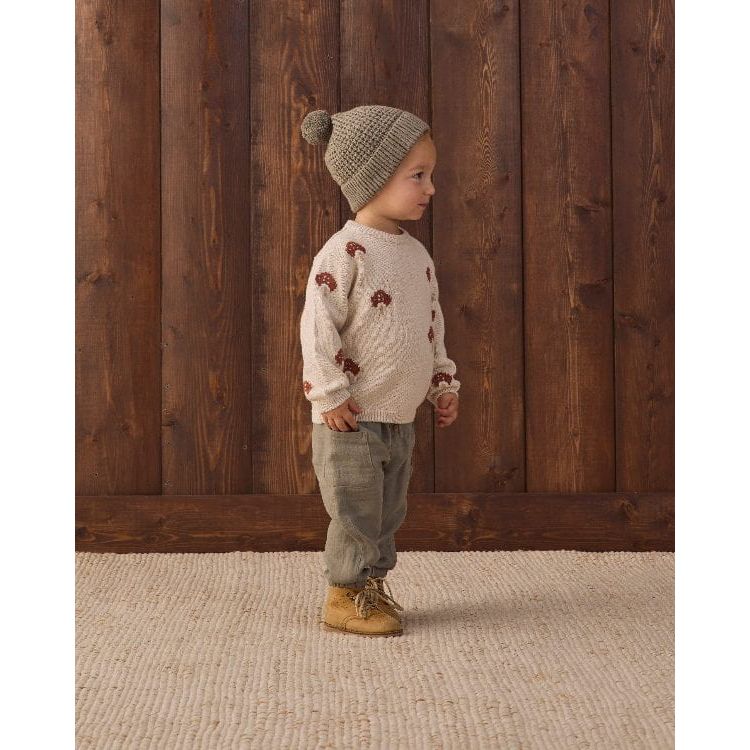 A toddler stands in profile wearing a cream knit sweater with brown bear motifs, a beige knit beanie with a pom-pom, olive pants, and tan boots, set against a dark wood plank wall and a light woven rug.