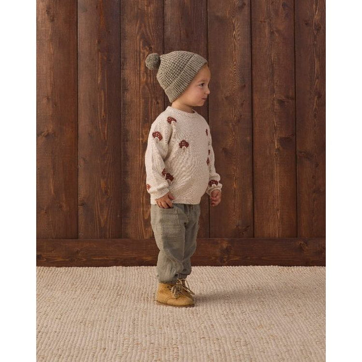 A toddler stands in profile wearing a cream knit sweater with brown bear motifs, a beige knit beanie with a pom-pom, olive pants, and tan boots, set against a dark wood plank wall and a light woven rug.