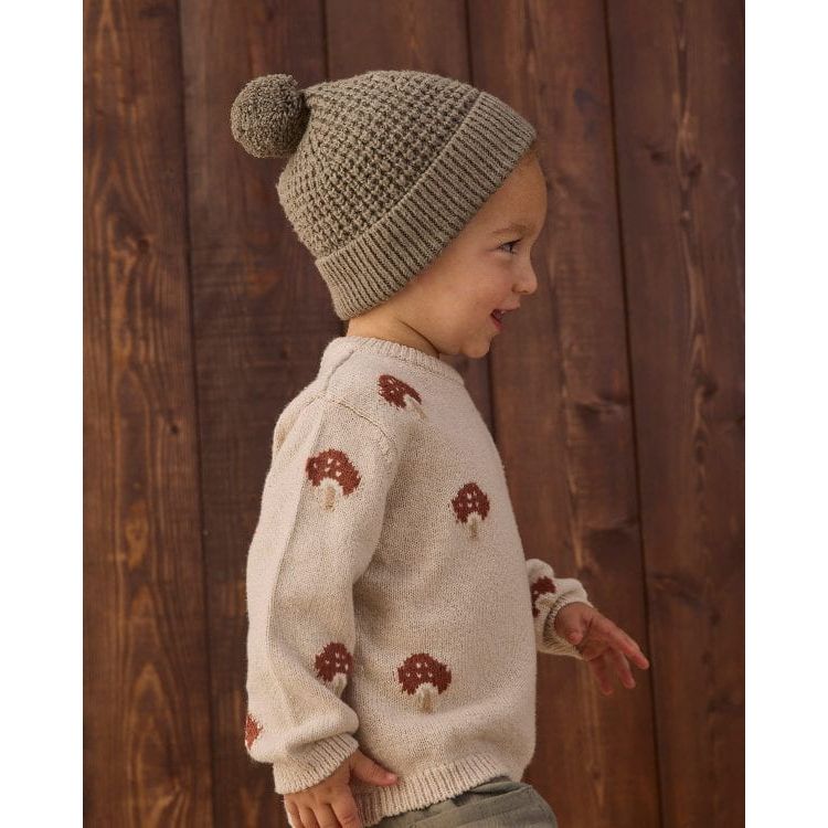 A toddler wearing a cream knit sweater with brown and white mushroom motifs, paired with a matching beige knit beanie, standing against a dark brown wooden plank background.
