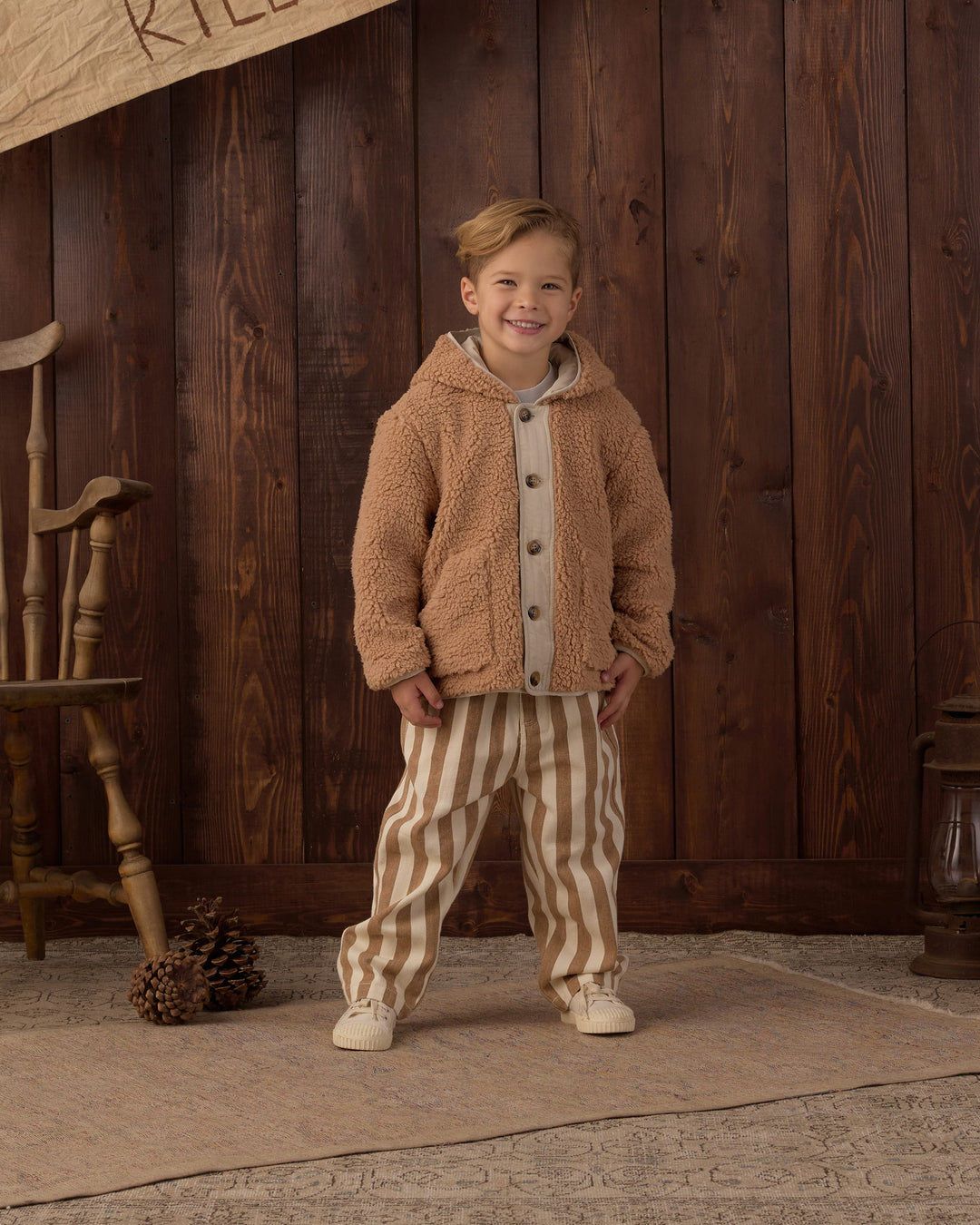 A young boy stands in front of a rustic dark wood plank wall wearing a tan sherpa fleece hooded button-up jacket over a light gray shirt and beige-and-white vertically striped pants.