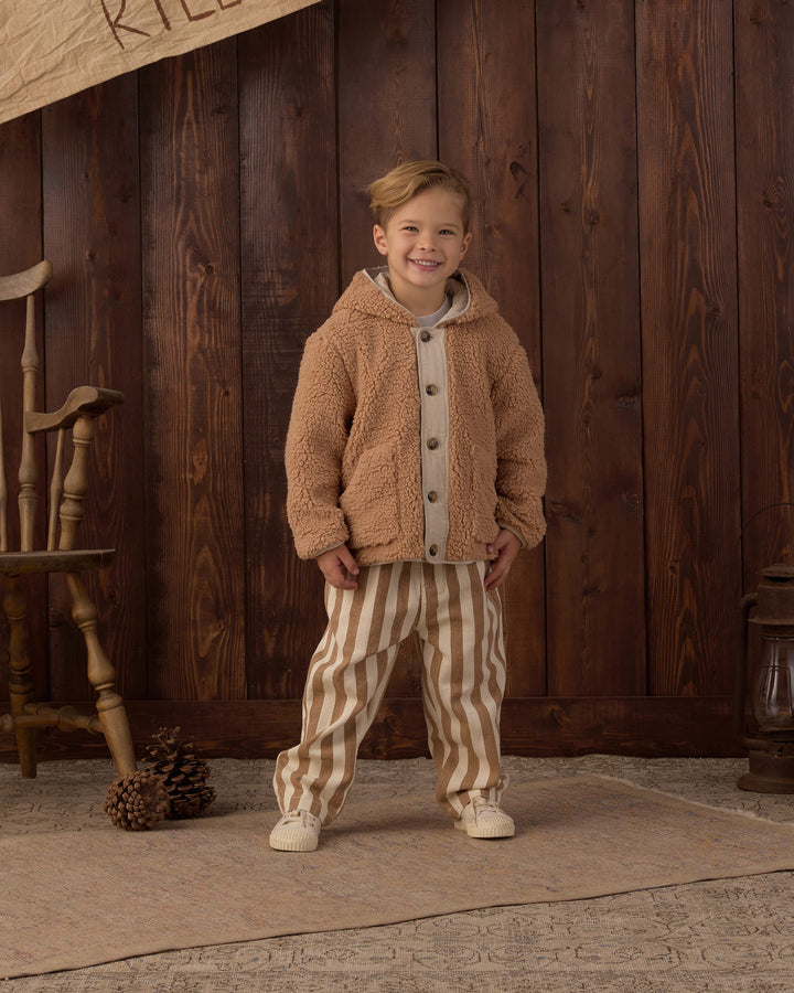 A young boy stands in front of a rustic dark wood plank wall wearing a tan sherpa fleece hooded button-up jacket over a light gray shirt and beige-and-white vertically striped pants.