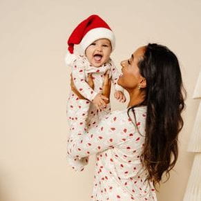 A woman and a baby wearing matching white Christmas-themed pajamas with red hearts, with the baby also wearing a red Santa hat. The woman is holding the baby up against a warm beige background.