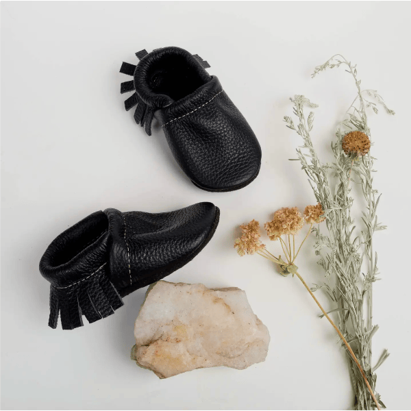 Pair of black leather baby shoes with fringes on a light gray background with a rock and dried plant.