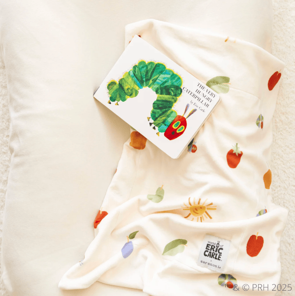 A baby blanket with a colorful print of the Green Monster from The Very Hungry Caterpillar book, styled on a white pillow with the book itself placed on top. The blanket features a white base with green leaves, red apples, and a green caterpillar.
