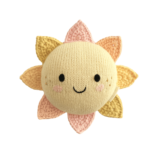 A cute, hand-knitted plush sun toy with a round cream-colored center, embroidered black eyes and smile, and soft pastel yellow and pink petals, isolated on a white background.