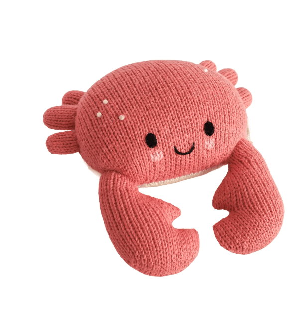 A knitted plush crab toy in a dusty coral pink color, featuring embroidered black eyes and a smiling mouth, with small white details on its shell, set against a plain white background.