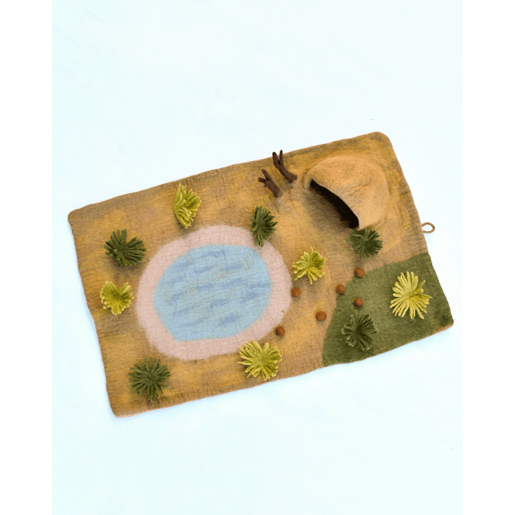 A handmade felted wool landscape rug featuring a light blue pond, green and yellow plants, and a brown cave, displayed on a clean white background.