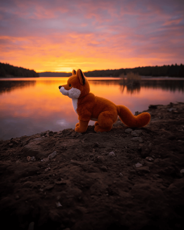 A plush fox toy sits on a dark rocky shoreline at sunset beside a calm lake, with orange and pink sky reflections in the water.
