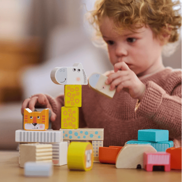 A young child with curly hair sits at a wooden table, playing with colorful animal and geometric wooden blocks. The child wears a dusty rose knit sweater and is captured in a warm, soft-focus indoor setting.