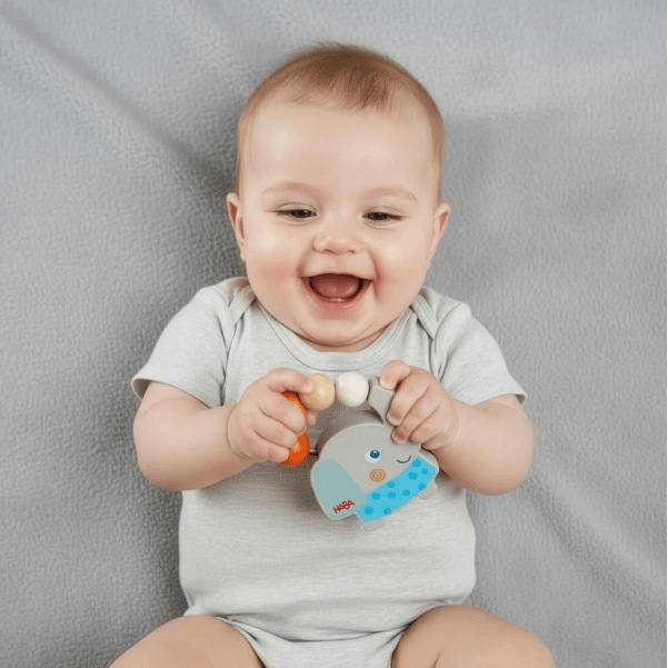 A baby wearing a light gray short-sleeve bodysuit holds a colorful elephant teething toy against a textured gray fabric background.