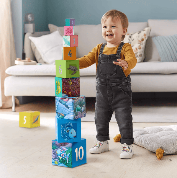 A toddler stands in a bright living room next to a tall stack of colorful children&