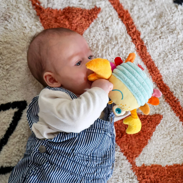 A baby wearing blue and white striped overalls and a white long-sleeve shirt lies on a textured cream and rust rug, biting and chewing on a soft plush toy with a light blue striped hat and yellow accents.