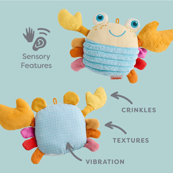 A plush crab sensory toy shown from two angles on a light blue background, with text and icons highlighting sensory features, crinkles, textures, and vibration. The toy has a cream face and belly, yellow-orange claws, a light blue striped textured body, and pink, orange, and red side accents.
