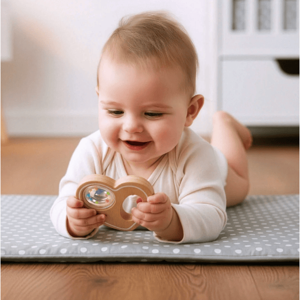 A baby lying on a gray and white polka dot play mat on a wooden floor, smiling while holding a wooden teething toy with a colorful mirror.