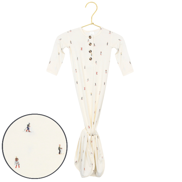 An off-white long-sleeve baby footie sleeper with a small skier pattern, hanging on a gold hanger with a circular inset showing a close-up of the print.