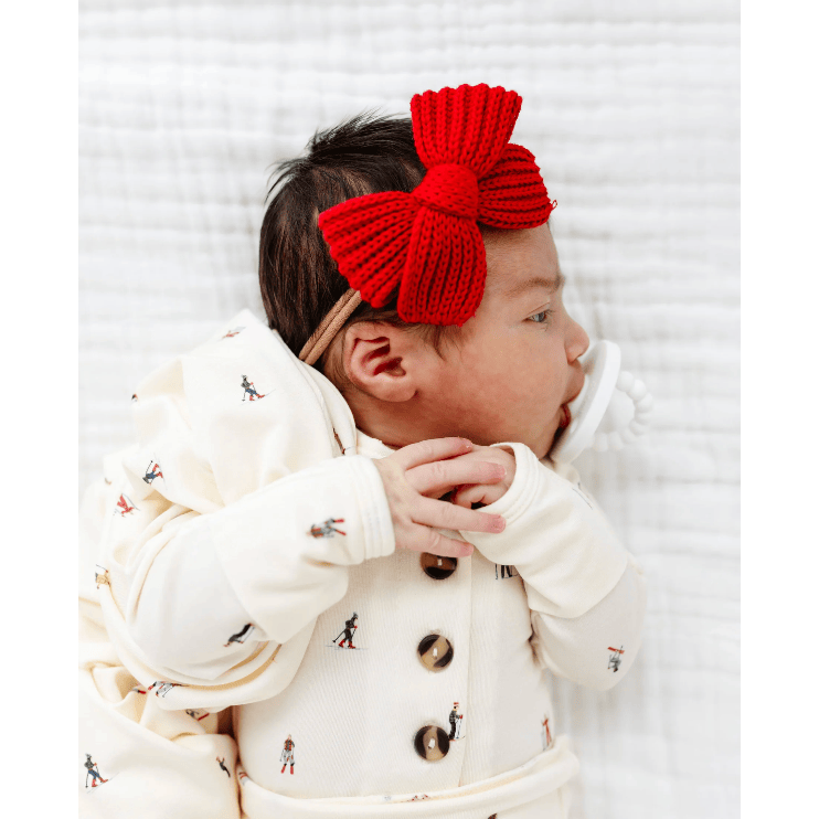 A baby wearing a cream-colored knit outfit with tiny printed figures and a large bright red knitted bow headband, lying on a white textured blanket.