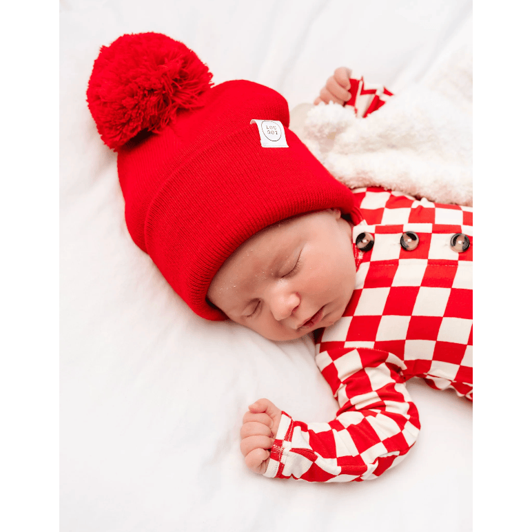 A sleeping newborn baby wearing a bright red knit beanie with a large pom-pom and a matching red-and-white checkered outfit, lying on a soft white blanket.
