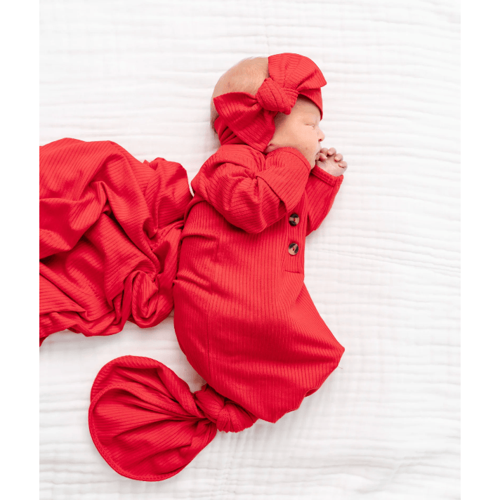 A sleeping newborn baby wearing a bright red ribbed long-sleeve bodysuit with a matching large bow headband and a matching red swaddle blanket, lying on a white textured blanket.