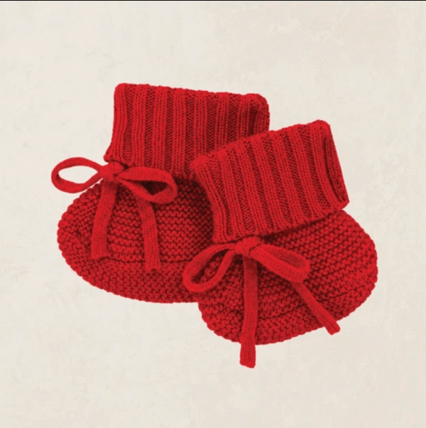 A pair of red hand-knit baby booties with ribbed cuffs and matching red bow ties, displayed on a textured off-white background.