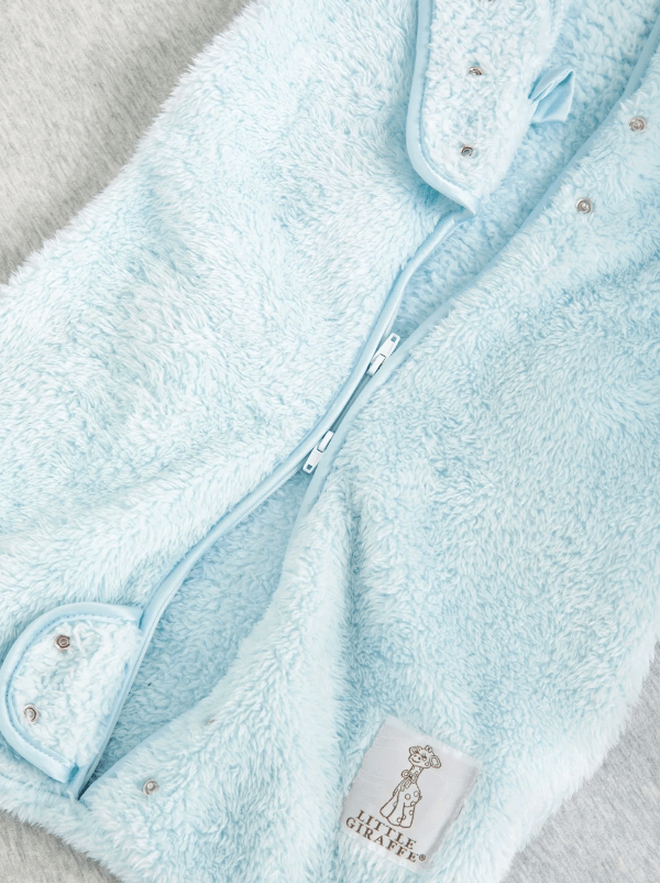 Close-up of a light blue baby outfit with a zipper and a brand label on a neutral background.