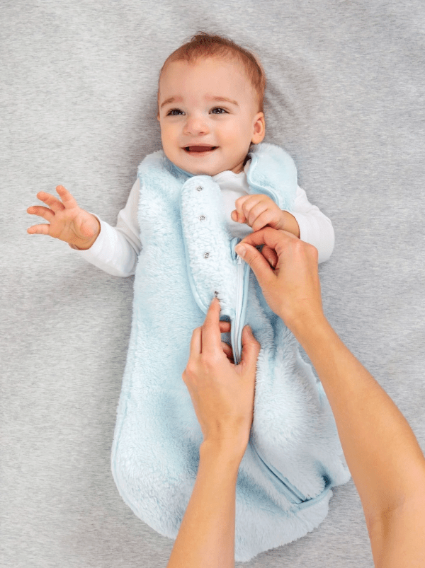 Baby lying on a gray blanket with a light blue swaddle, being held by an adult's hands.