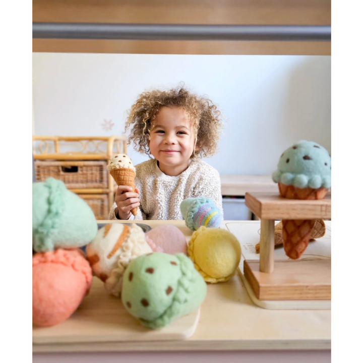 Child holding a toy ice cream cone with colorful play food items on a table.