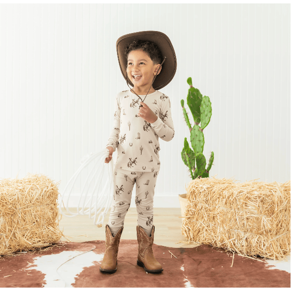 Child wearing a cowboy hat and boots in a rustic setting with hay bales and a cactus.
