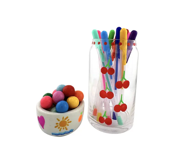 Colorful straw set with cherry design in a glass container next to a small bowl of multicolored balls on a white background.