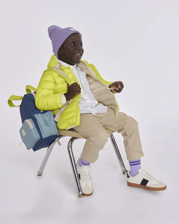 A young child wearing a bright lime green puffer jacket, beige pants, white sneakers, and a lavender beanie sits on a metal chair with a blue and lavender backpack against a plain light gray studio background.