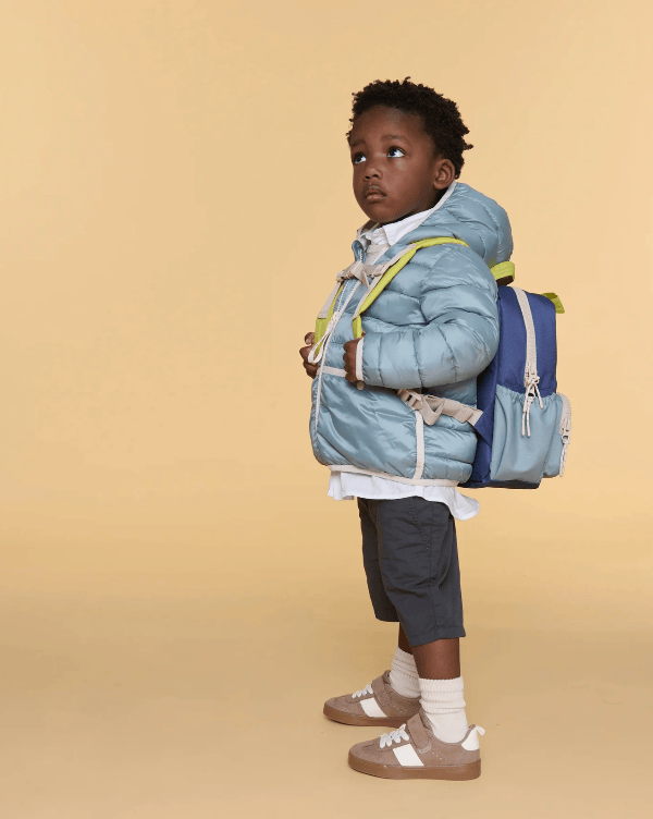 A toddler stands in profile wearing a light blue puffer jacket, dark gray shorts, white socks, and tan shoes, with a blue and yellow backpack. The photo is shot against a solid warm beige studio background.