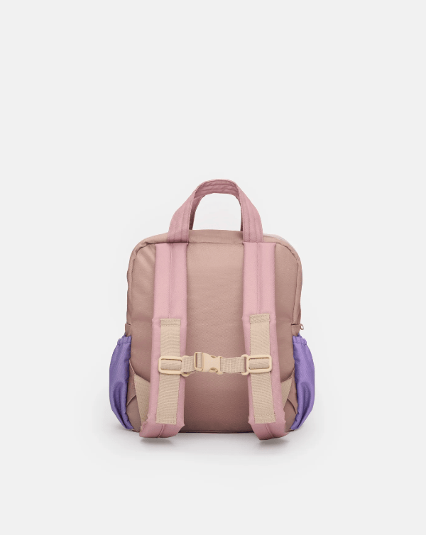 A small backpack shown from the back, featuring a dusty pink body with lavender purple side pockets and matching pink straps with beige plastic buckle details, set against a plain light gray background.