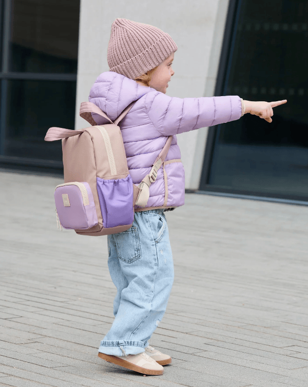 A toddler wearing a light purple puffer jacket, light blue jeans, and a pink ribbed beanie, carrying a pastel purple and beige backpack while pointing outdoors.