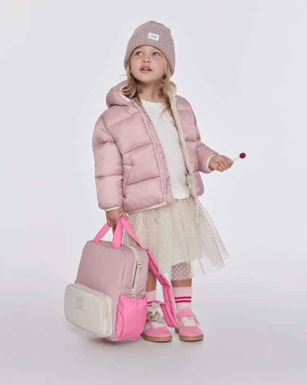 A young girl stands against a light gray studio background wearing a pink puffer jacket, cream top, sheer beige skirt, pink shoes, and a pink beanie, while holding a pink and cream backpack and a small red object in one hand.