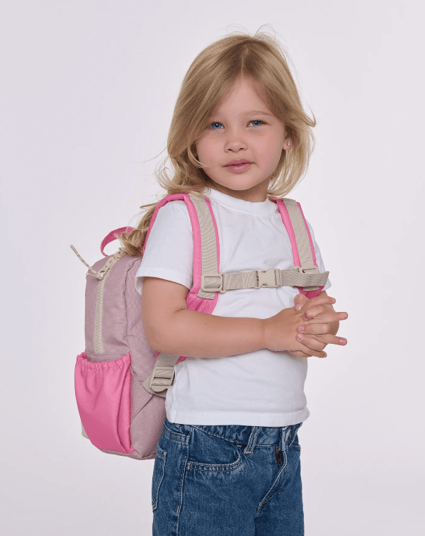 A young blonde girl wearing a white t-shirt and blue jeans, carrying a pink and beige backpack over her shoulder, photographed against a solid light gray background.