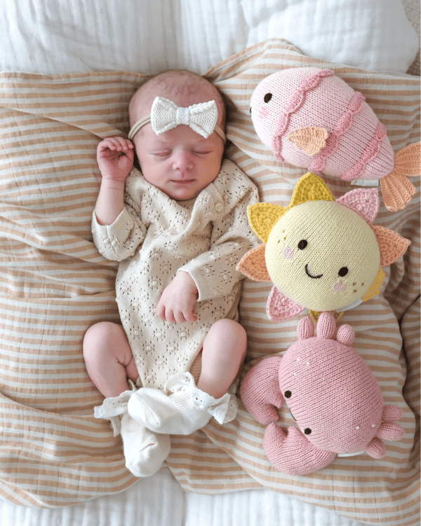 A sleeping newborn baby lies on a striped beige and white blanket, surrounded by handmade pink and cream crochet toys including a fish, sun, and crab, with a matching cream knit bodysuit and bow headband.