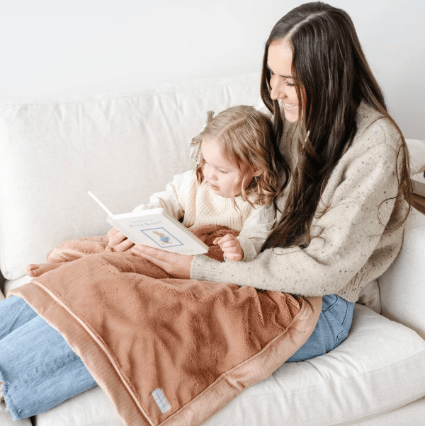 A woman and a young girl sit on a white sofa reading a book, with a soft brown blanket draped across both of them. The woman wears a cream speckled sweater and blue jeans, and the child wears a cream knit top.