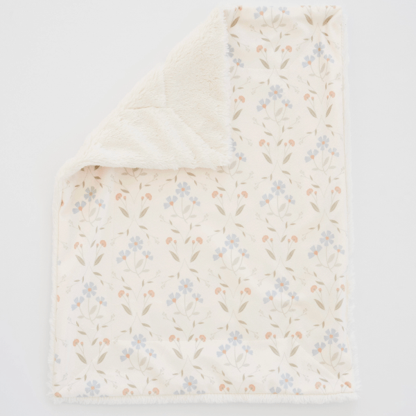 A soft baby blanket with a plush cream sherpa lining, featuring a cream fabric printed with a delicate pattern of small blue flowers, peach blossoms, and green leaves. The blanket is shown with one corner folded over to reveal the soft cream sherpa side, set against a clean white background.