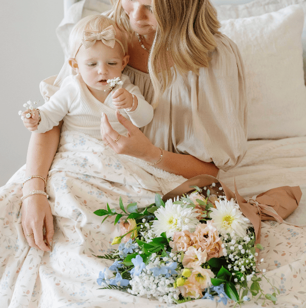 A woman and a young child sit together on a bed, surrounded by a floral bouquet. The woman wears a soft beige blouse, and the child wears a white long-sleeve dress with a matching beige bow headband.